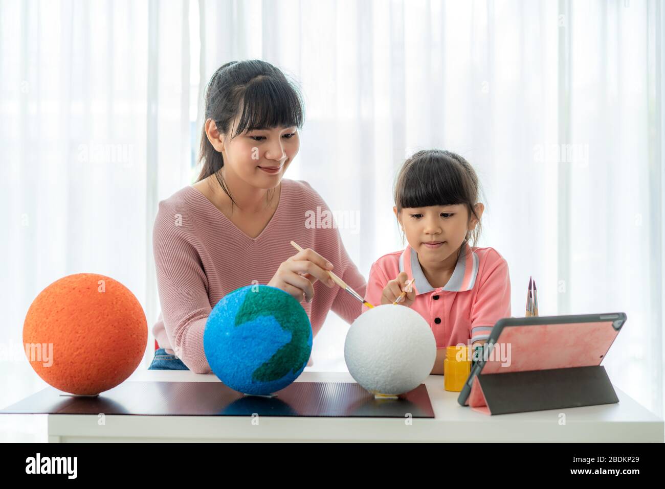 Asian elementary schoolgirl with mother painting the moon in science class learning about the solar system via video conference with teacher and other Stock Photo