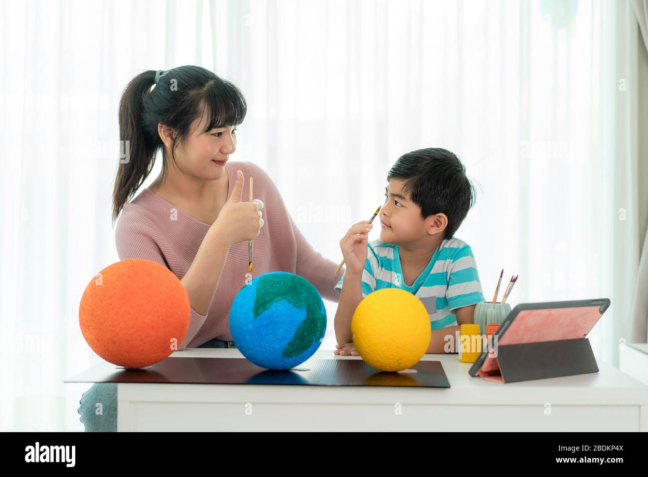 Asian elementary schoolboy with mother painting the moon in science class learning about the solar system via video conference with teacher and other Stock Photo