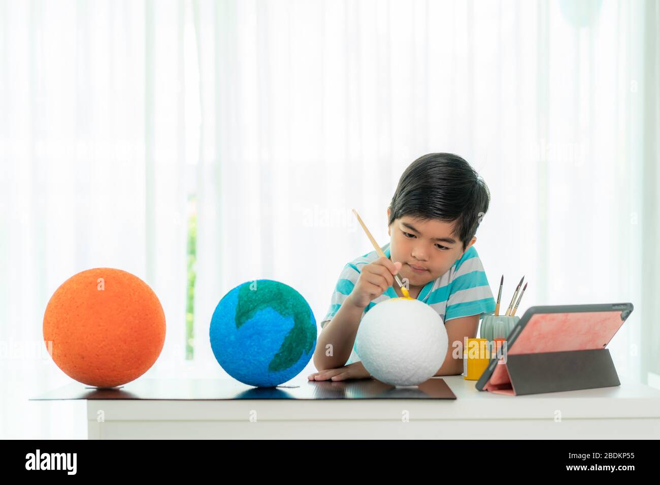 Asian elementary schoolboy painting the moon in science class learning about the solar system via video conference with teacher and other classmates a Stock Photo