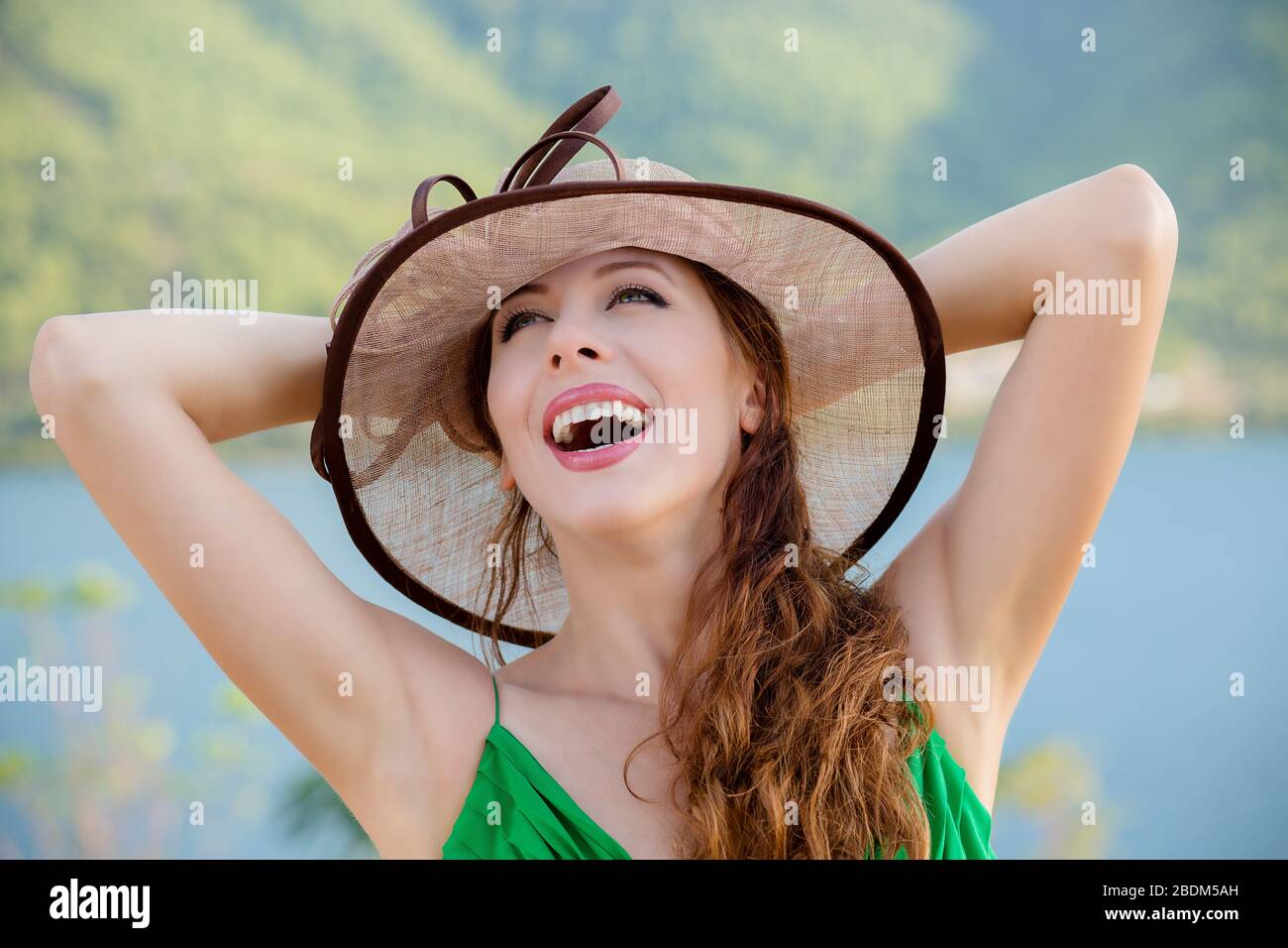 Closeup portrait happy young pretty woman in grey shirt looking upwards, hands on head in air relaxing, isolated park, lake on background. Lady in hat Stock Photo