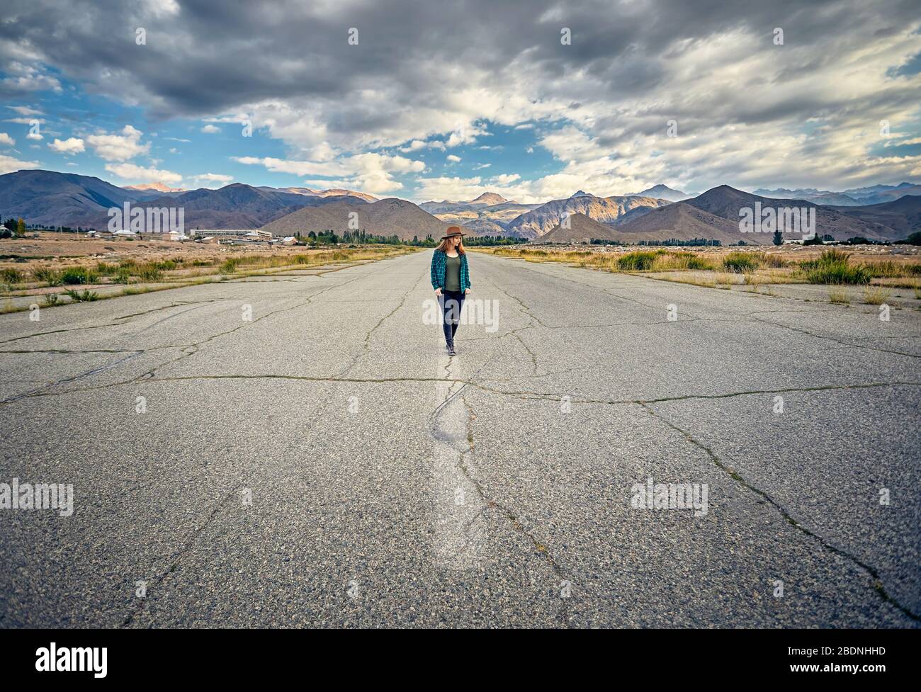 Woman in hat and checked shirt walking on the wide asphalt road with mountains and cloudy sky background Stock Photo