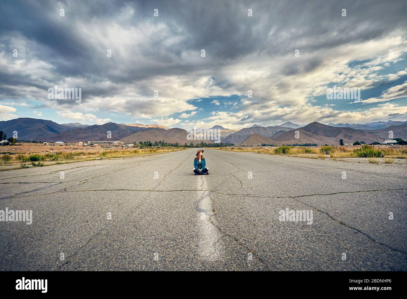 Woman in hat and checked shirt sitting with cross leg on the wide asphalt road with mountains and cloudy sky background Stock Photo