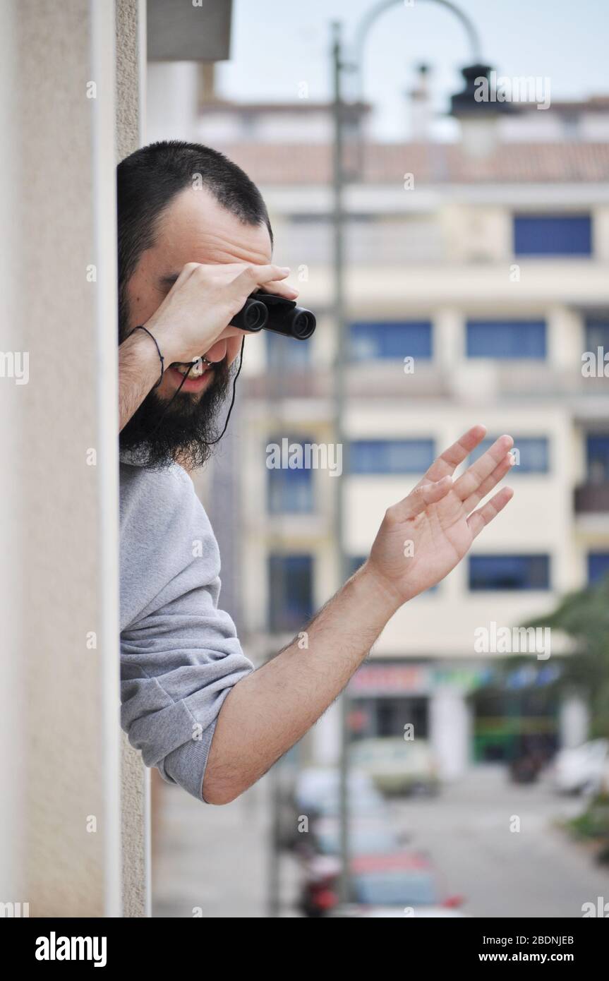 Young man waving family while he is leaning out of condominium window looking through binoculars. Man watching through window with binoculars in Spani Stock Photo