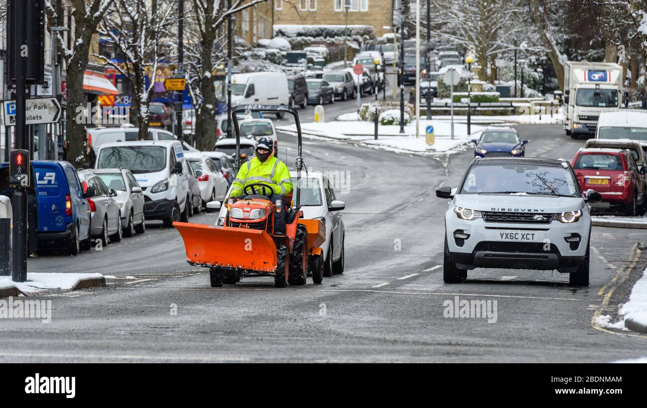 Winter snow in town (busy road, cars parked, compact tractor waiting at town centre traffic lights) - Brook Street, Ilkley, Yorkshire, England, UK. Stock Photo
