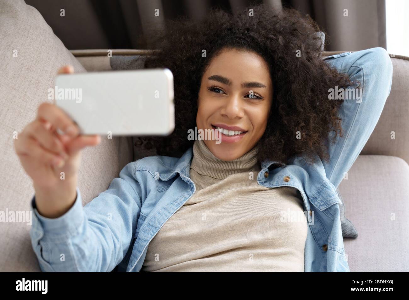 Relaxed african woman hold smartphone watching movie, making video call at home. Stock Photo