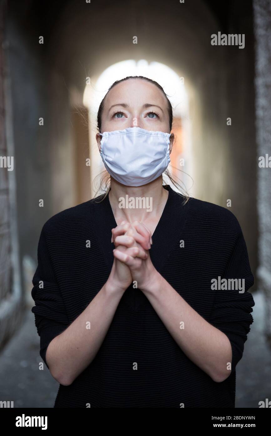 Coronavirus outbreak. Young caucasian woman wearing medical protection face mask praying over coronavirus global pandemic, for salvation of humanity Stock Photo