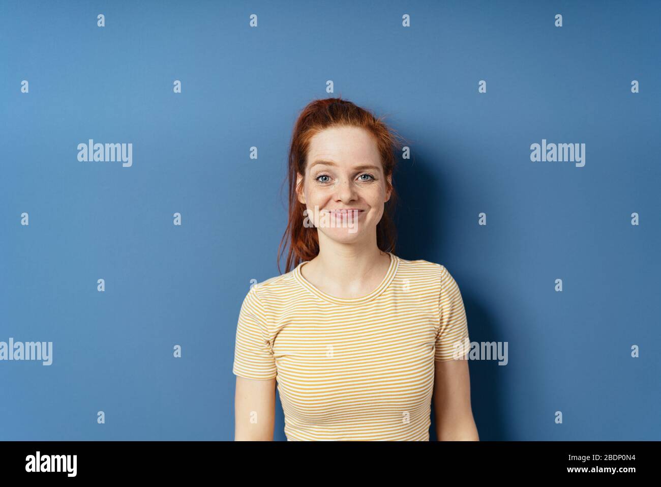 Pert young woman with a gleeful grin looking at the camera on a blue studio background with copy space Stock Photo