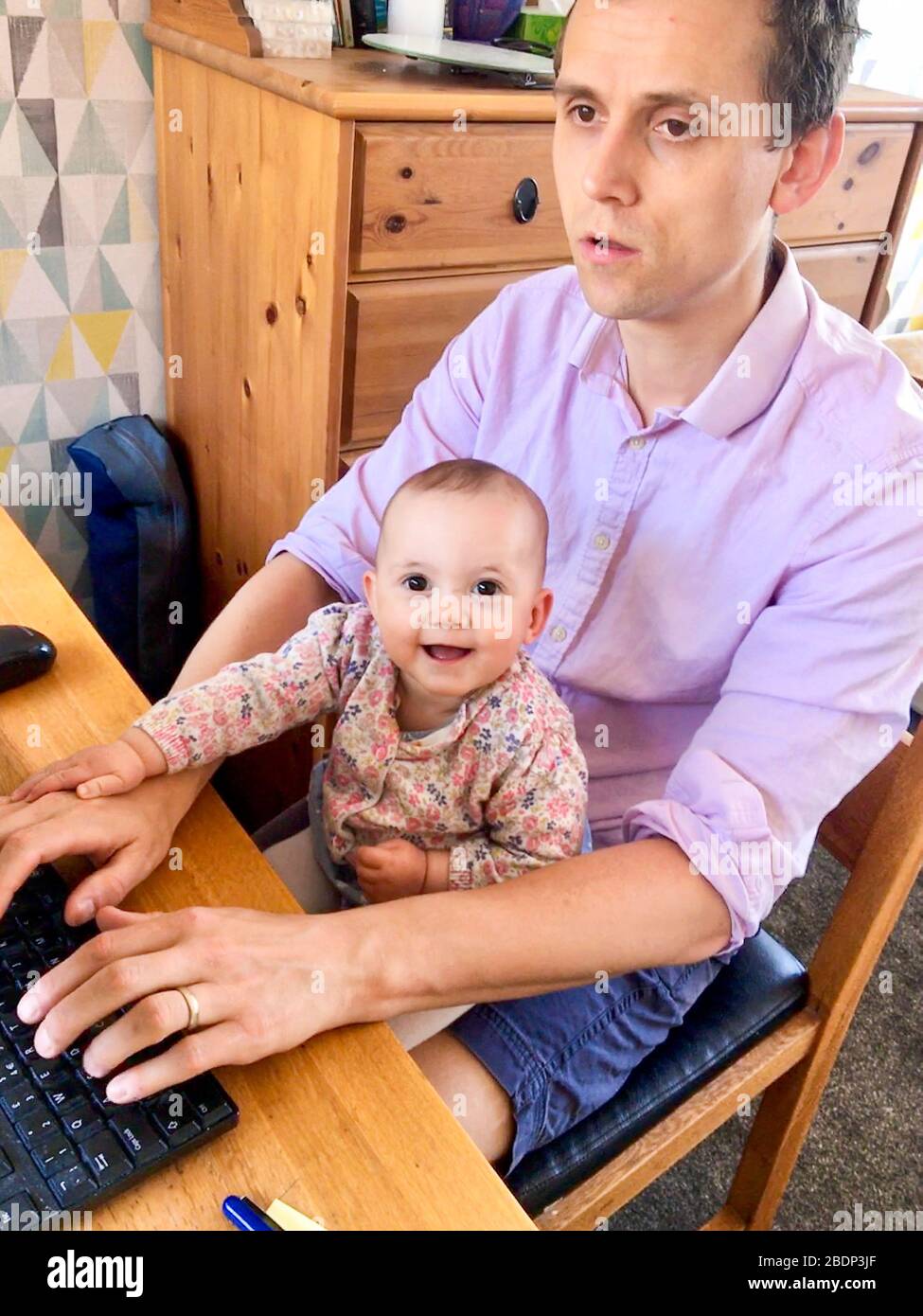 A higher education employee working from home WFH during the Covid 19 pandemic assisted by his seven month old daughter. England UK Stock Photo