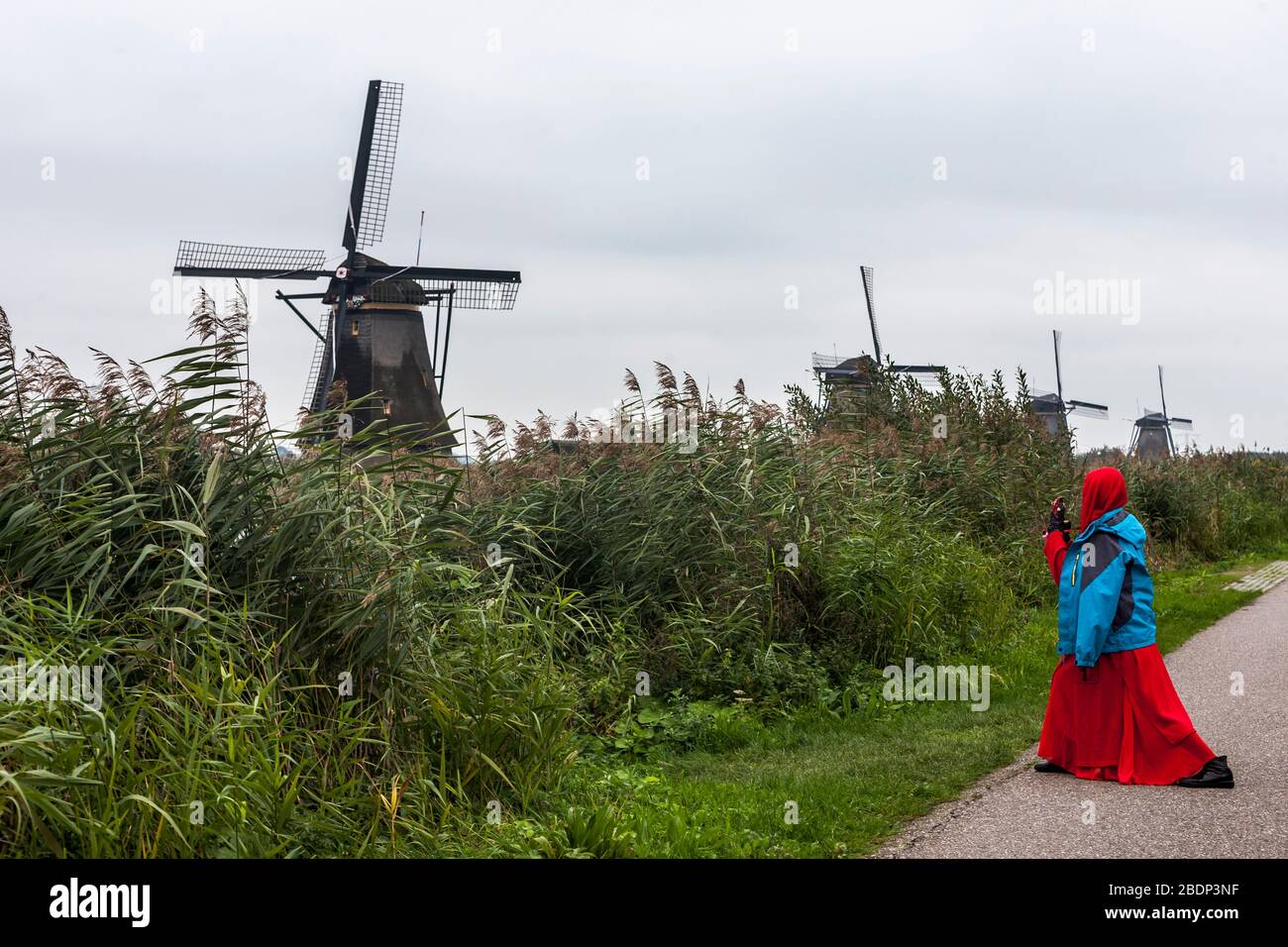 Girl in bright red headscarf and dress photographs the Overwaard windmills, Kindersdijk UNESCO World Heritage Site, South Holland, Netherlands Stock Photo