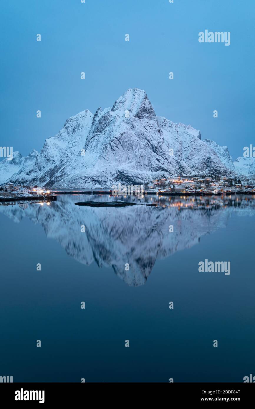 Lonely mountain perfectly reflected in cold, calm waters of northern fjord. Snow covered mountain ridge over early morning calm bay of Reine, Norway Stock Photo