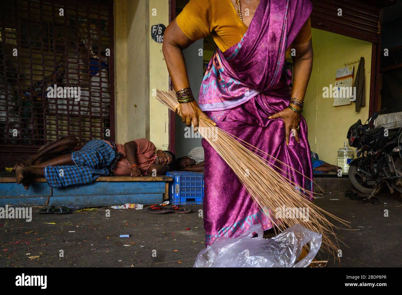 Indian woman sweeping as men in the background sleep, Koyambedu Market,  Chennai, India Stock Photo