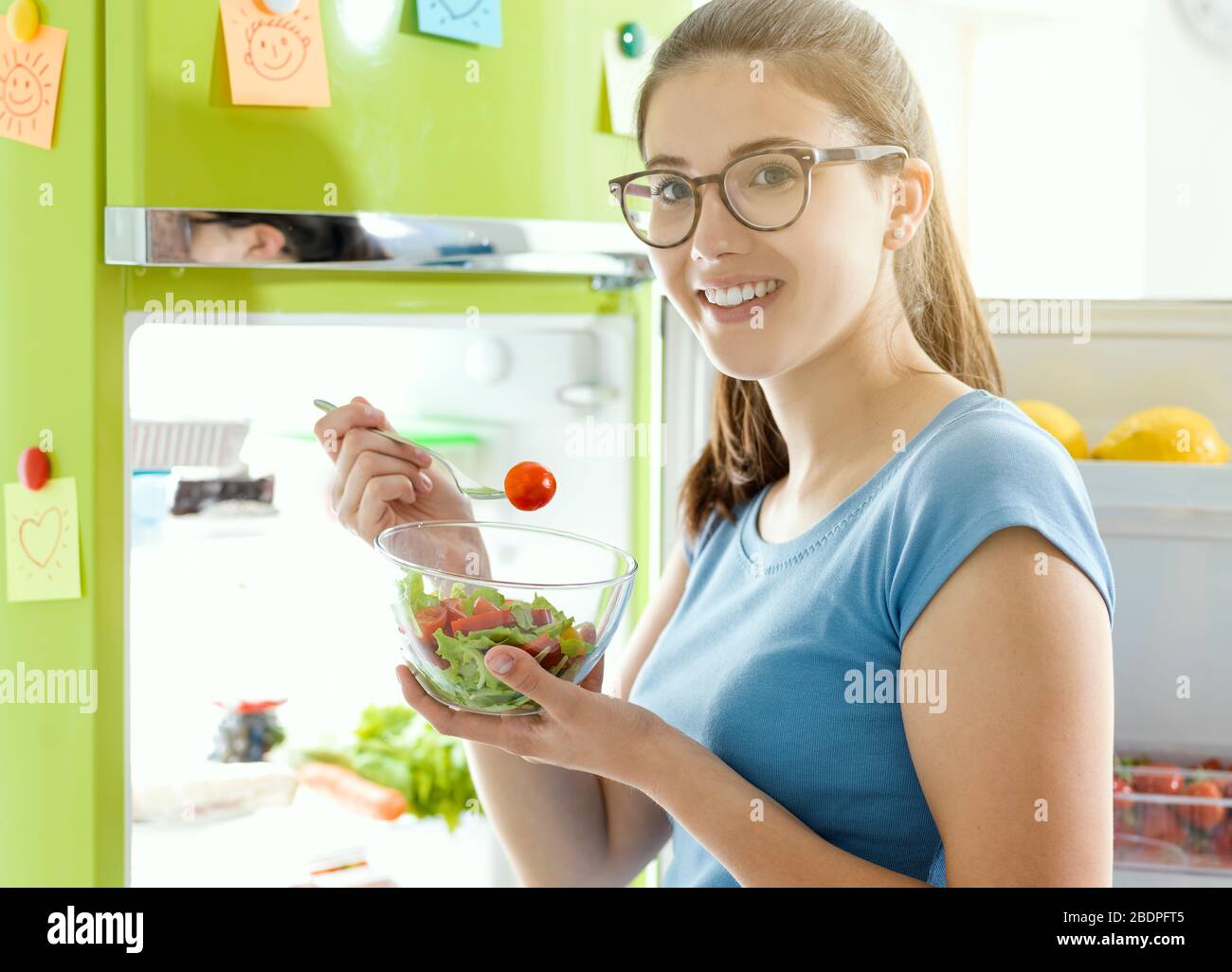 Smiling woman eating some fresh salad next to the fridge, healthy vegan food and dieting concept Stock Photo