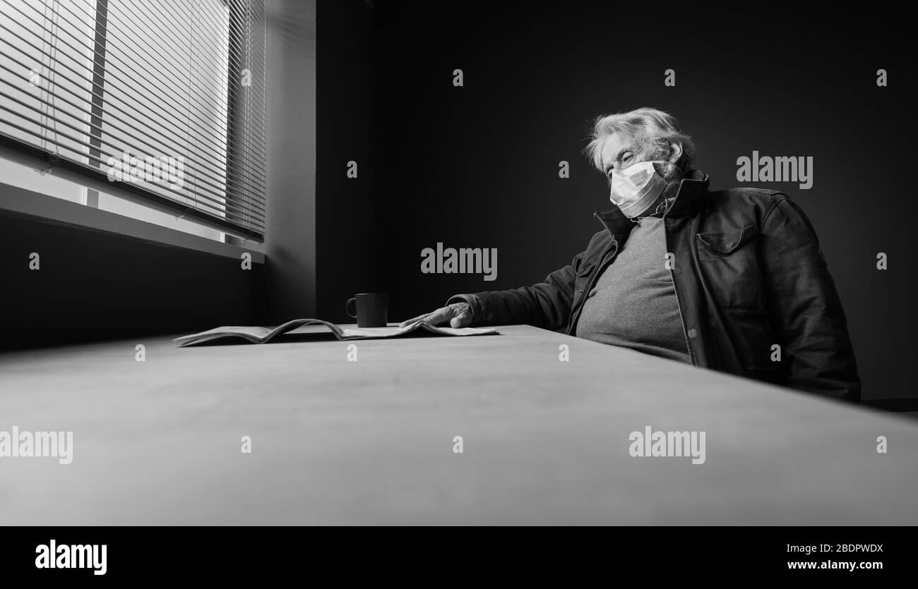Lonely senior sitting at home alone, wearing a face mask and reading a newspaper, virus covid-19 emergency measures and elderly safety concept Stock Photo