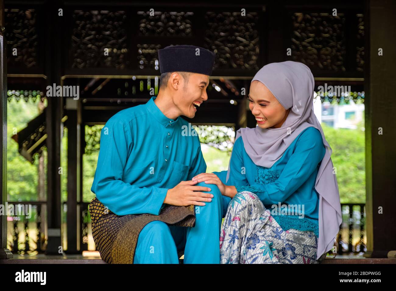 Young couple of malay muslim in traditional costume having happy conversation during Aidilfitri celebration at traditional wooden house. Raya and Musl Stock Photo