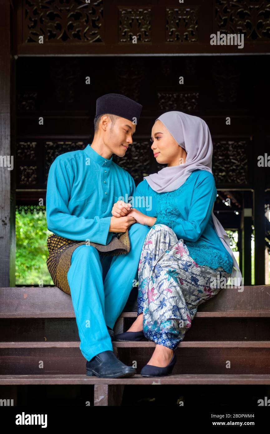 A portrait of young couple of malay muslim looking each other in traditional costume during Aidilfitri celebration at the traditional wooden house. Ra Stock Photo
