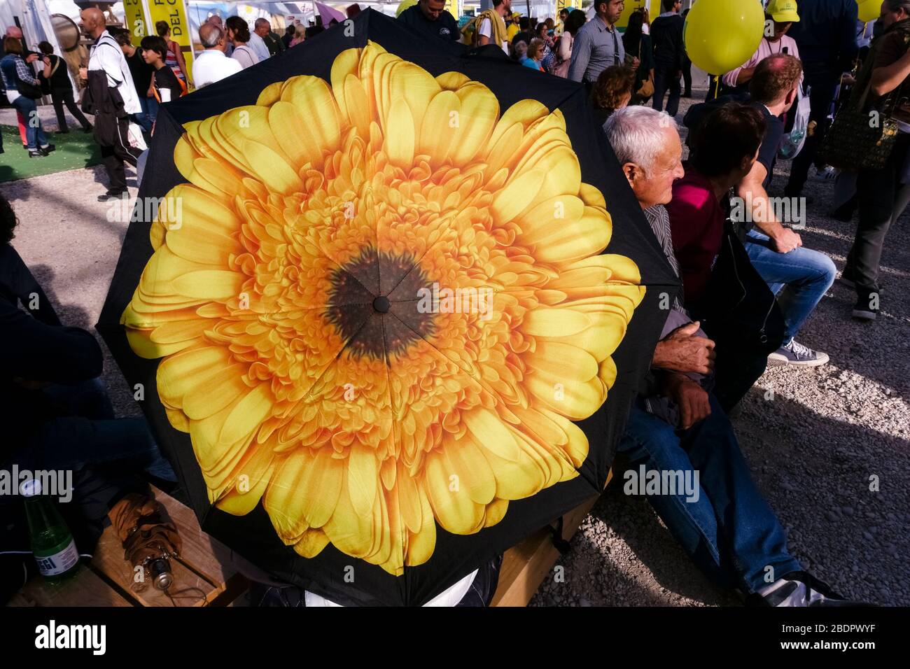 Elderly man sitting in the crowd in a sunny day. Parasol umbrella with a large yellow, orange flower, in the foreground. Stock Photo