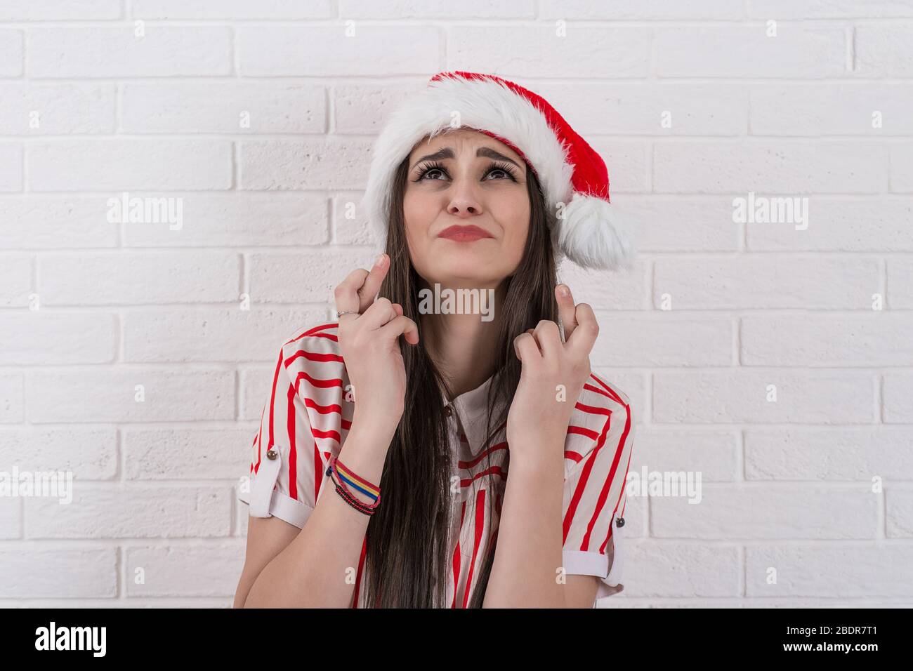 Closeup portrait of beautiful young woman wearing red santa claus hat crossing fingers, closing eyes, wishing praying for a miracle, isolated on white Stock Photo