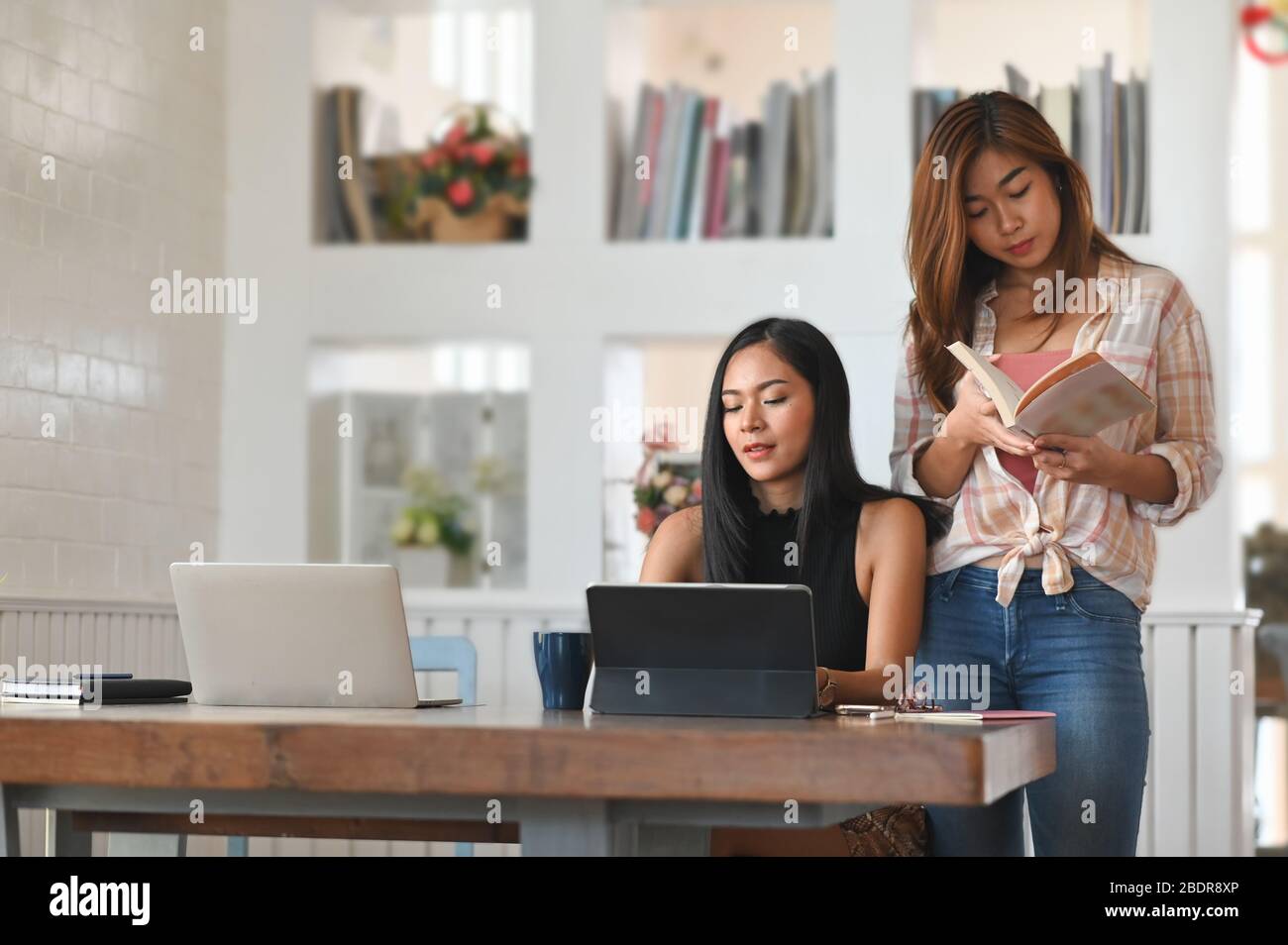 Photo of two beautiful woman while tutoring a study lesson from university at the modern wooden table with computer tablet, laptop and books with comf Stock Photo