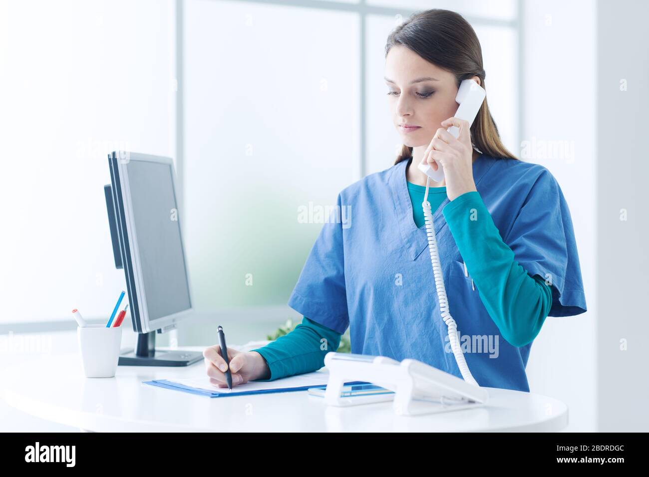 Young female doctor and practitioner working at the reception desk, she is answering phone calls and scheduling appointments Stock Photo