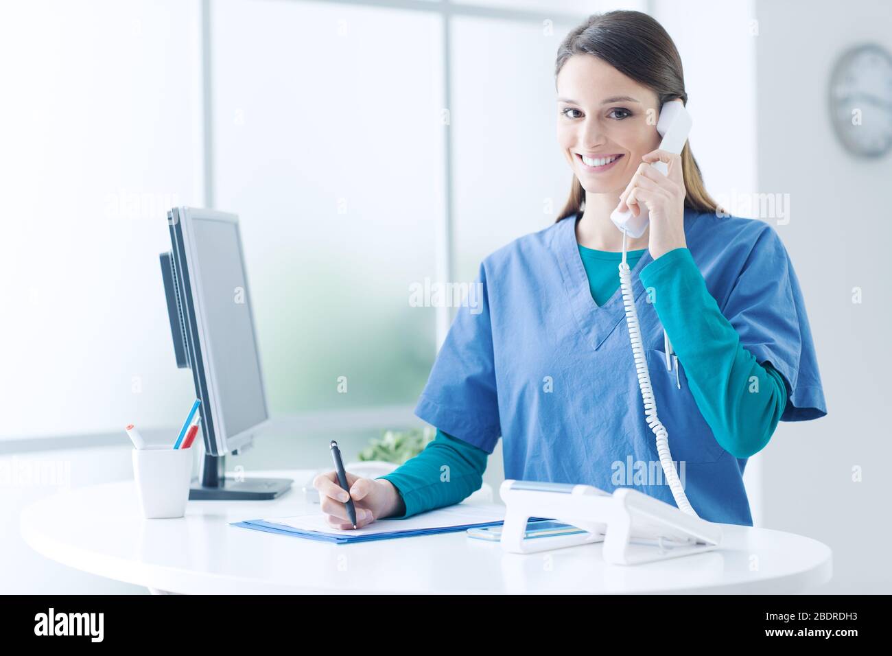 Young female doctor and practitioner working at the reception desk, she is answering phone calls and scheduling appointments Stock Photo