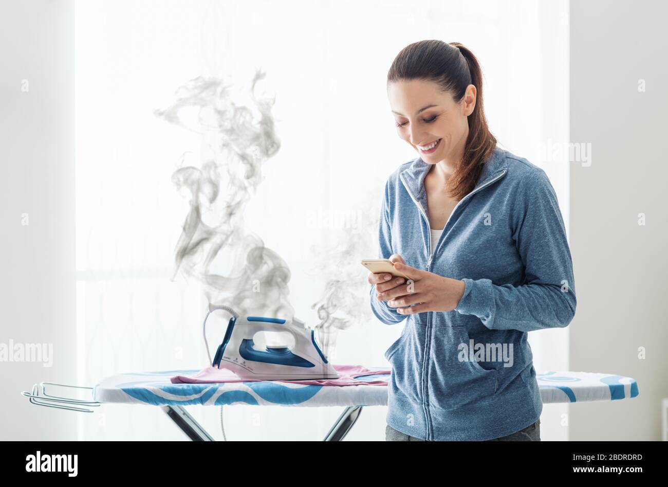 Careless smiling housewife at home texting with her smartphone, she has left the iron on the ironing board Stock Photo