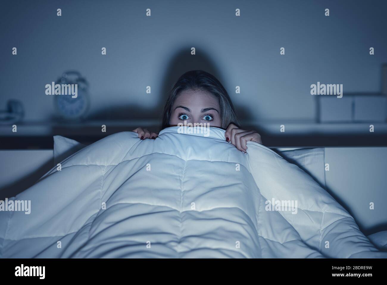 Woman watching a scary horror movie on tv late at night, she is frightened and hiding under the blanket Stock Photo