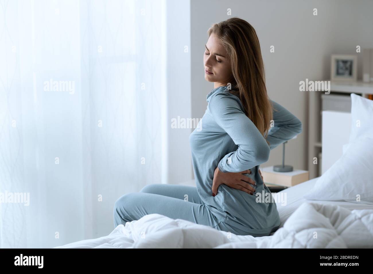 Young woman with back ache, she is sitting on the bed and touching her