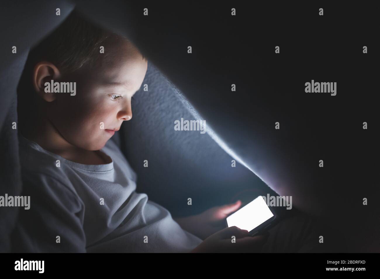 A child under a blanket playing with a smartphone, sitting on the Internet. The concept of spending time in safe isolation. Stock Photo