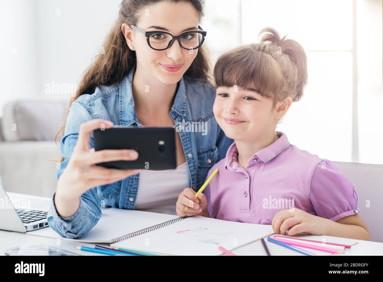 Cute girl and her young mother taking selfies with a smartphone at home, they are smiling and having fun Stock Photo