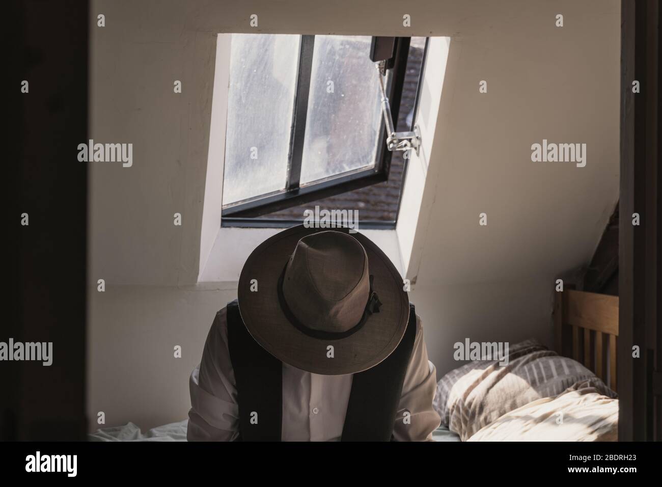A man looking down wearing a fedora hat and waistcoat sitting on a bed with a window behind. With a desaturated, classic edit Stock Photo