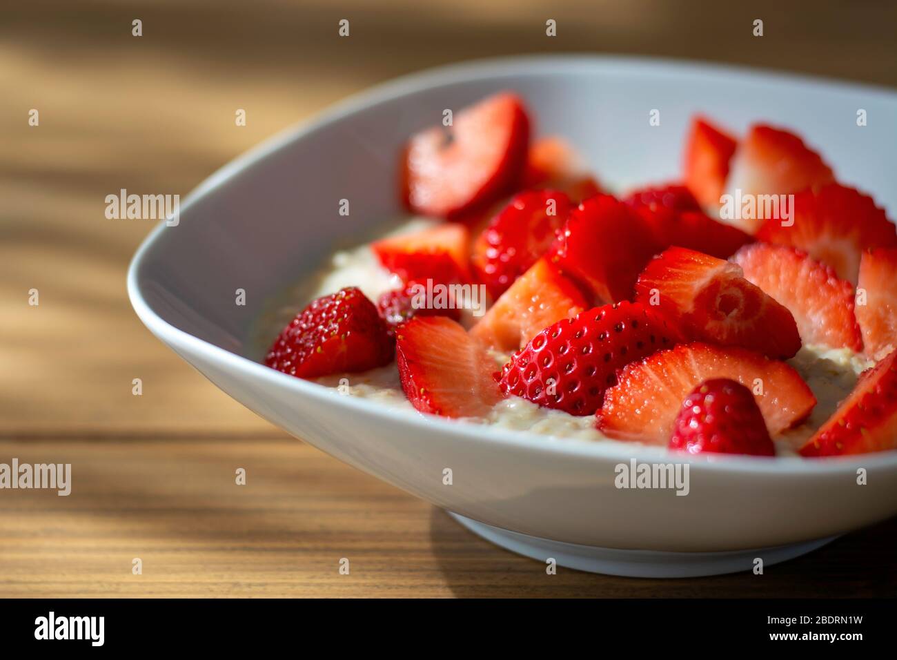 Homemade oatmeal porridge bowl with sliced strawberries on wooden table in morning sunlight. Healthy meal. Porridge oats Stock Photo