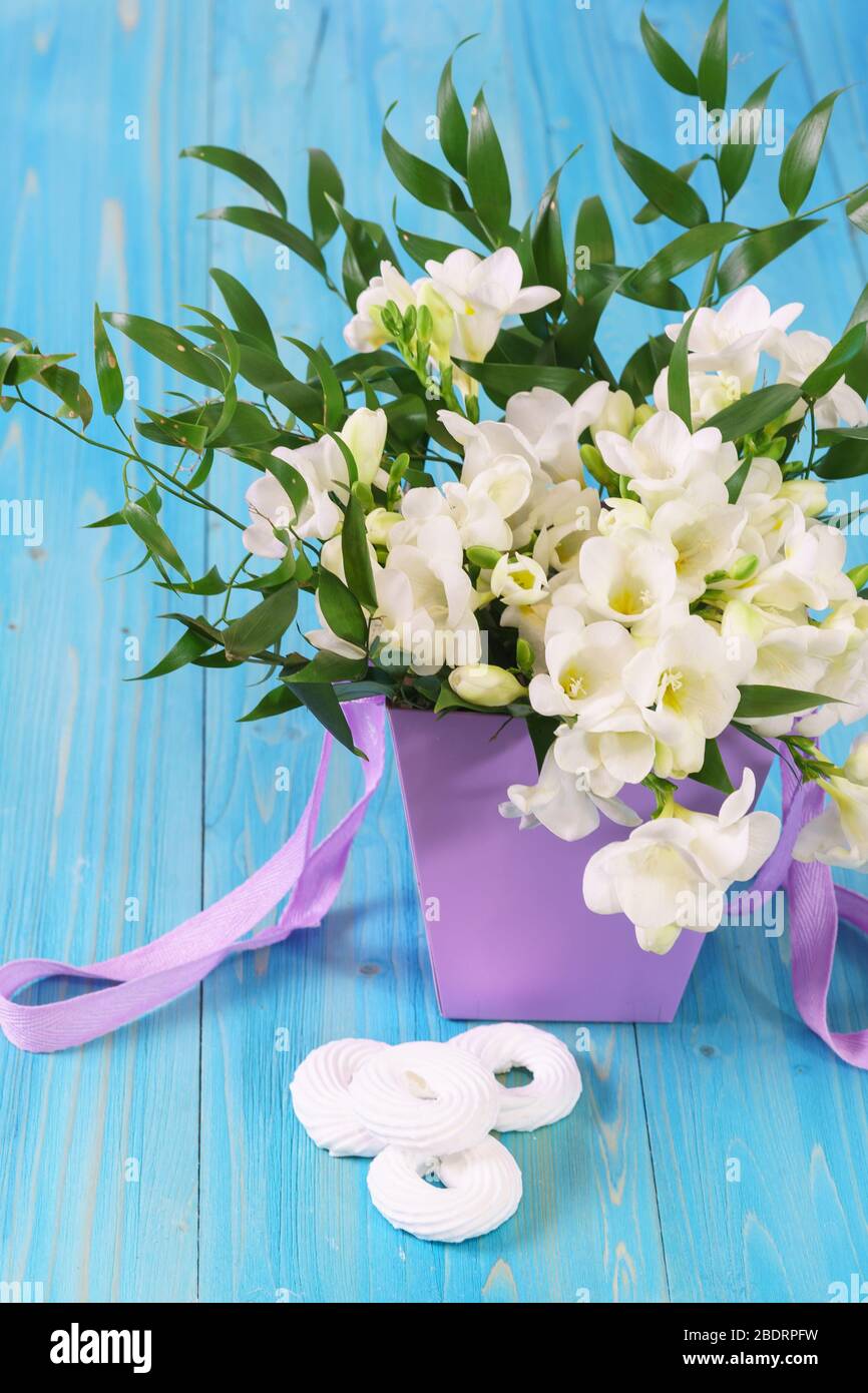 A delicate bouquet of white freesia in a lilac basket and a few airy meringue cakes on a blue wooden table. Spring card Stock Photo