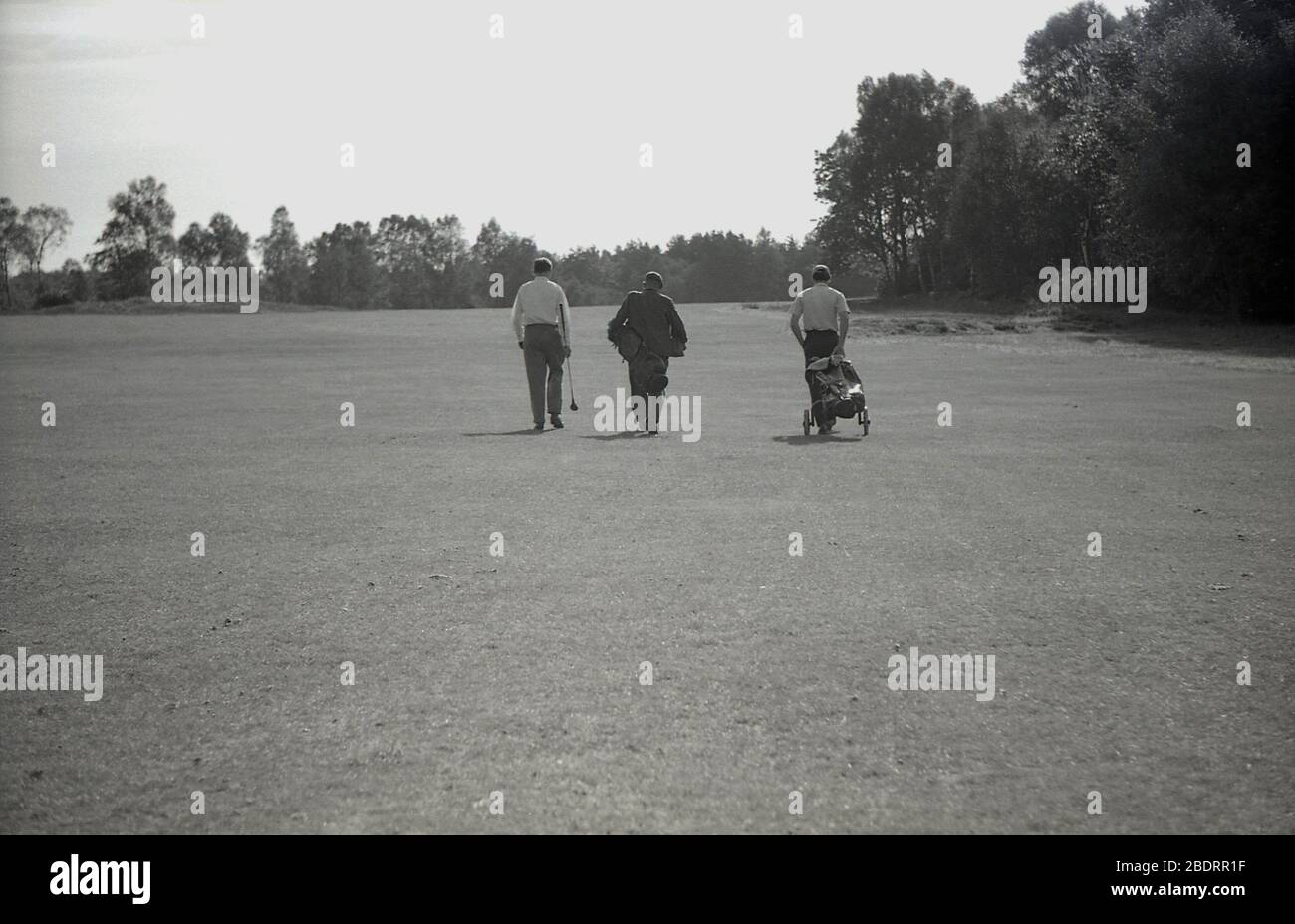 1960s, historical, on a golf course, in a line, three men walking down a fairway, two golfers and a caddie who is carrying clubs for one of them  and who is wearing a suit, while the other golfer is pulling a golf bag on a trolley of the era. Stock Photo
