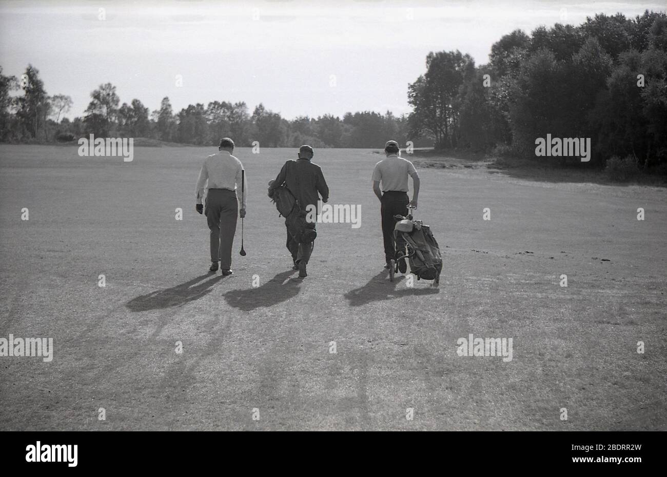 1960s, historical, on a golf course, in a line, three men walking down a fairway, two golfers and a caddie who is carrying clubs for one of them  and who is wearing a suit, while the other golfer is pulling a golf bag on a trolley of the era. Stock Photo