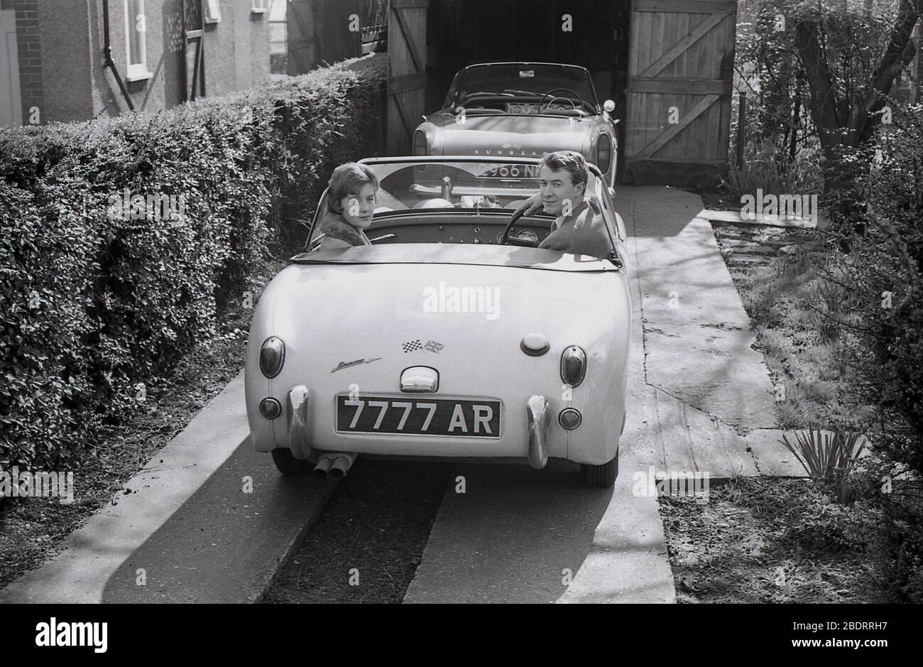 1960s, historical, a young adult couple sitting in an Austin Sprite sports car parked beside a hedge on a narrow concrete driveway, Eastholm Green, England, UK. A Sunbeam Alpine sportscar is also on the drive, parked half inside a wooden framed garage. Stock Photo