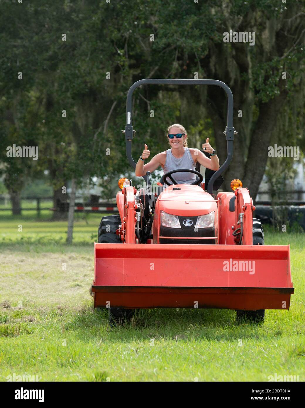 Blond woman driving a tractor mowing a pasture.  The image is straight on.  She is smiling and giving two thumbs up sign. Stock Photo