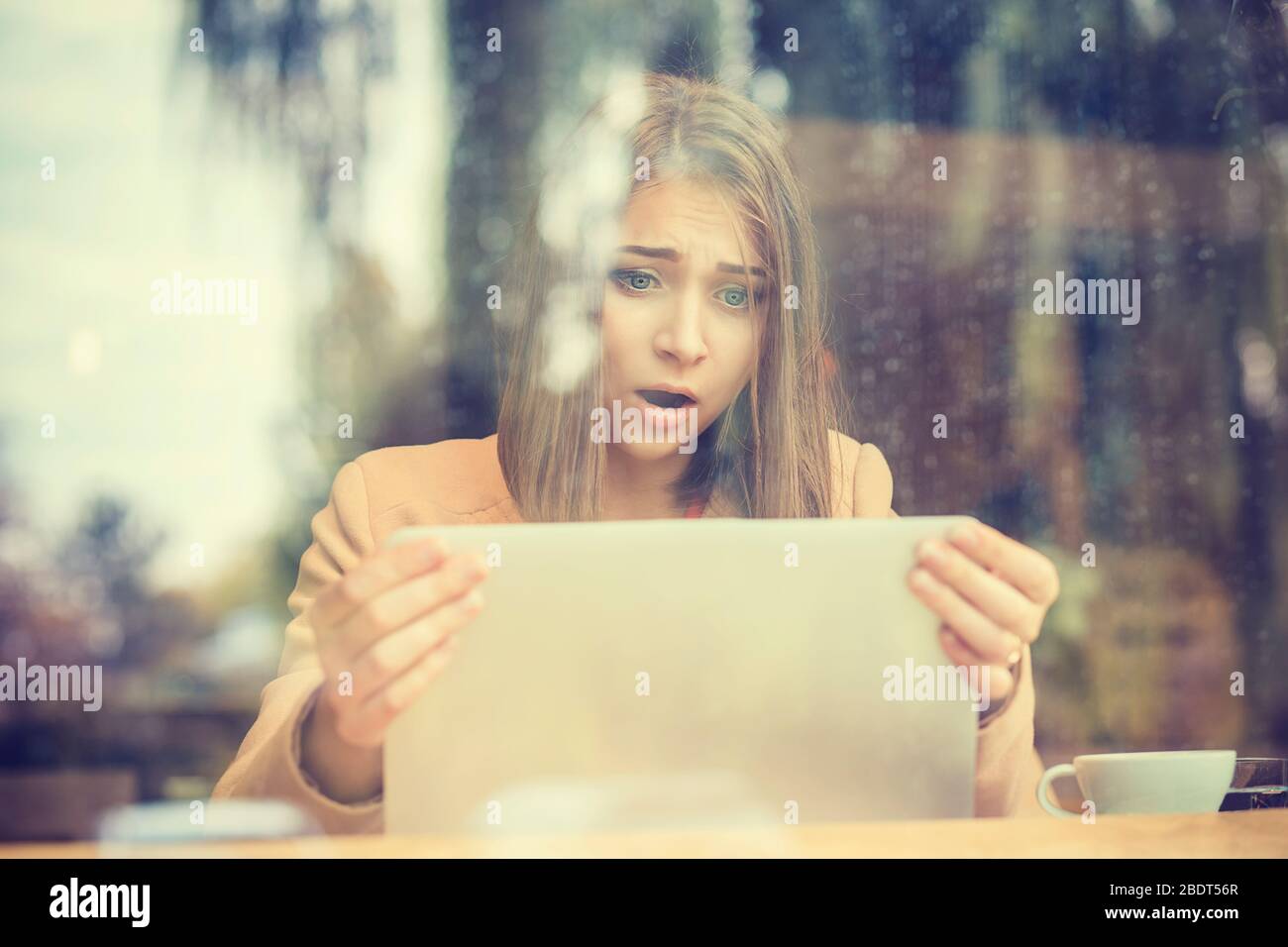 Shocked woman holding computer, laptop tablet screen looking surprised in coffee shop Stock Photo