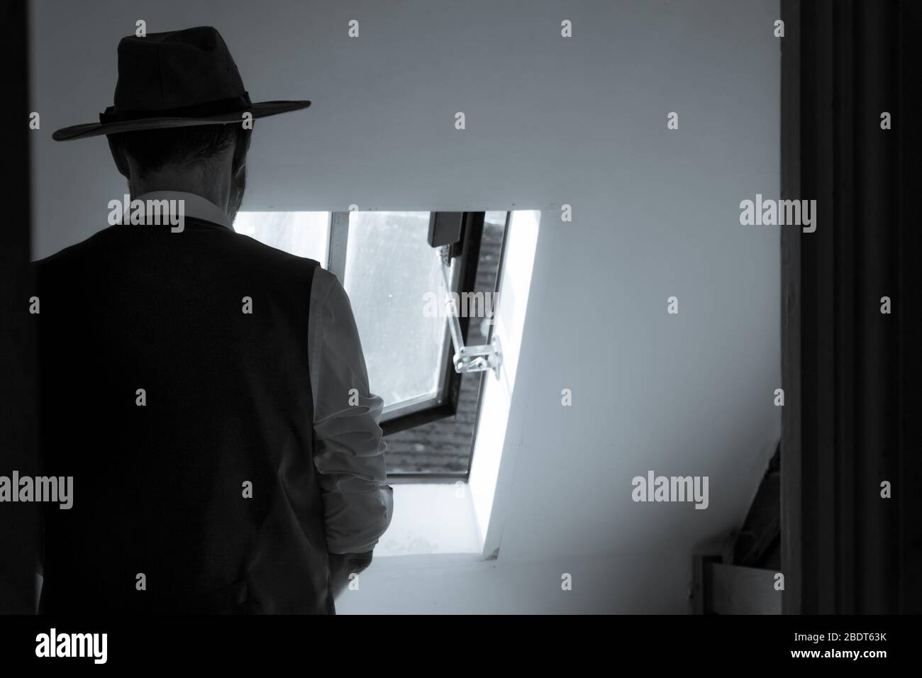 A man looking out of a window, back to camera, wearing a waistcoat and fedora hat. With a classic black and white edit Stock Photo