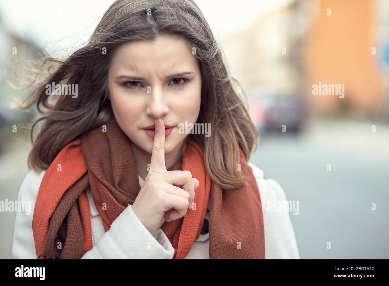 Shh Woman wide eyed asking for silence or secrecy with finger on lips hush hand gesture cityscape outdoor background Pretty girl placing fingers on li Stock Photo
