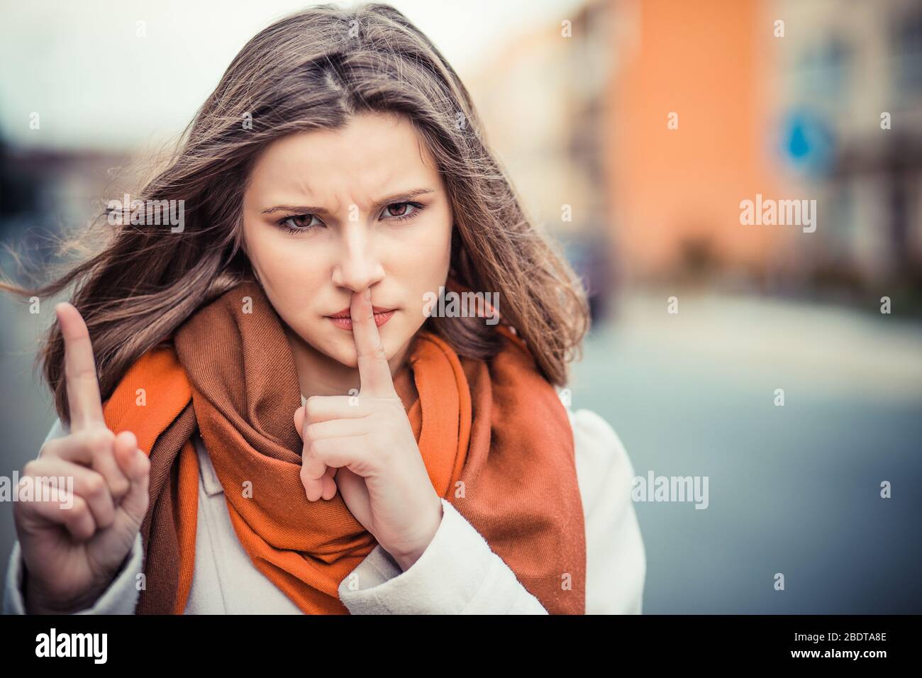 Woman asking for silence or secrecy with finger on lips hush hand gesture and woman wagging her other finger city background wall.  Shhh sign symbol. Stock Photo