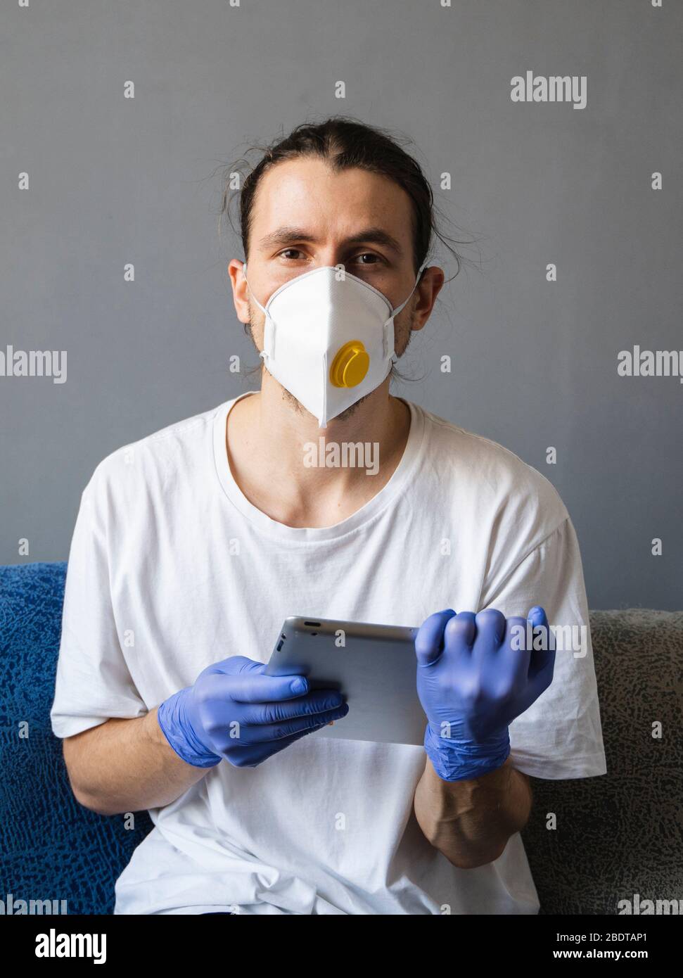 Man in white shirt, medical mask and rubber gloves sits at home and works with tablet on a sofa during quarantine. Man, designer, artist, architect Stock Photo