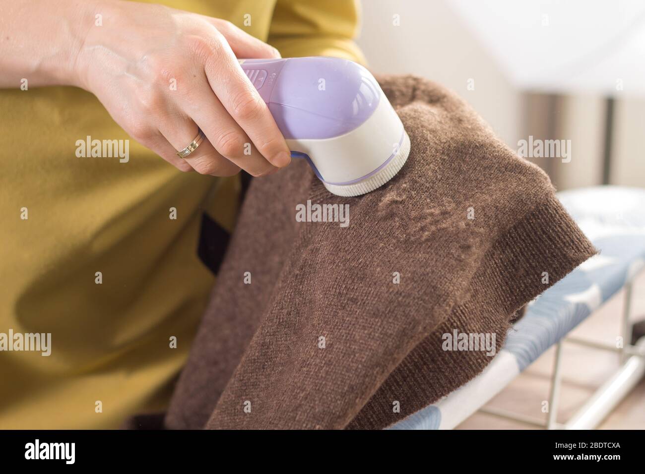The woman holds an electric shaver in her hand to clean mottled clothes and cleans the sleeve of a brown wool sweater. Stock Photo