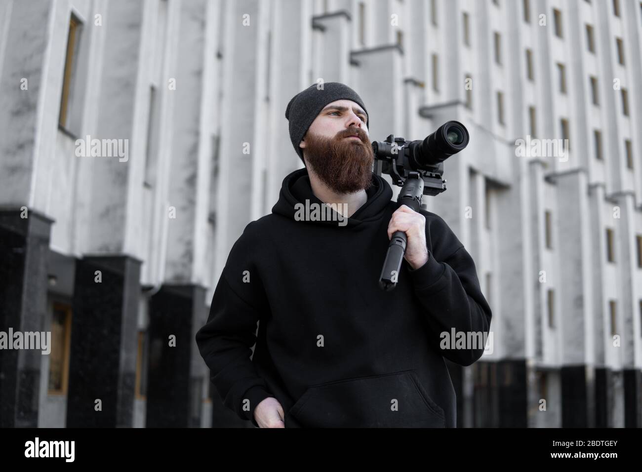 Bearded Professional videographer in black hoodie holding professional camera on 3-axis gimbal stabilizer. Filmmaker making a great video with a Stock Photo