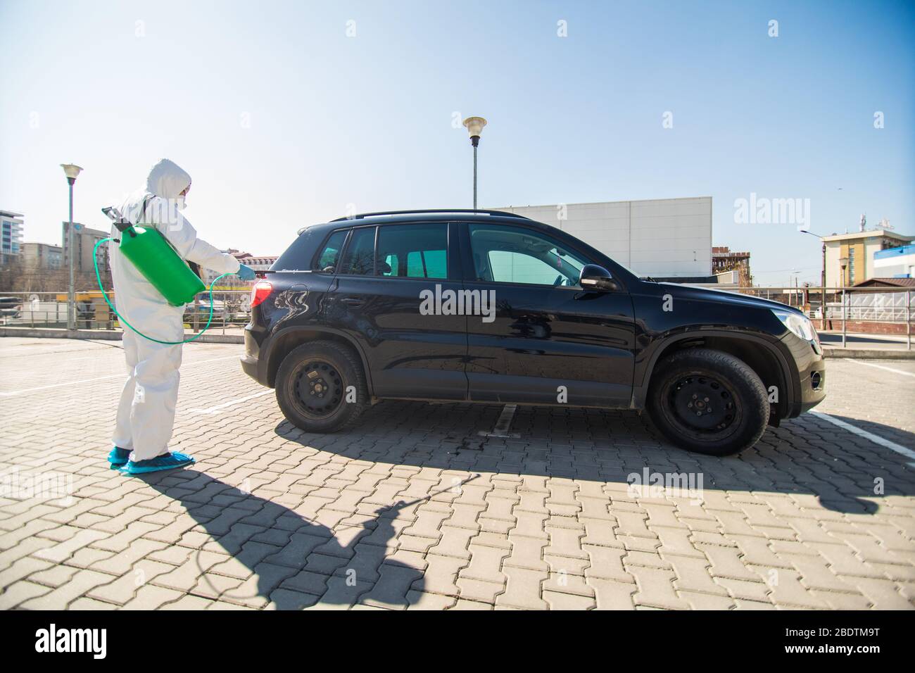 Disinfector in a protective suit and mask sprays disinfectants of car outdoors. Coronavirus Pandemic. Stock Photo