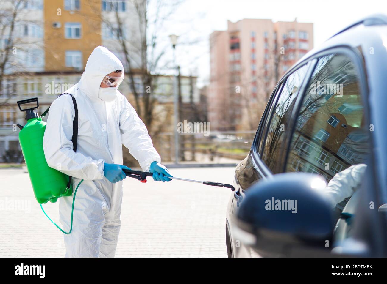 Disinfector in a protective suit and mask sprays disinfectants of car outdoors. Coronavirus Pandemic. Stock Photo