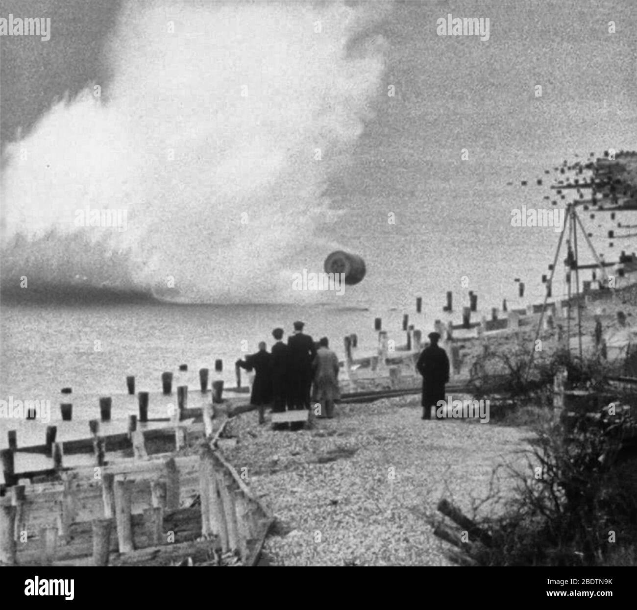 Movie still, showing an inert, practice version of the Upkeep bouncing bomb being dropped during a training flight by members of RAF 617 Squadron at Reculver bombing range, Kent. The bomb's designer, Barnes Wallis, and others watch the practice bomb strike the shoreline. 1943 Stock Photo
