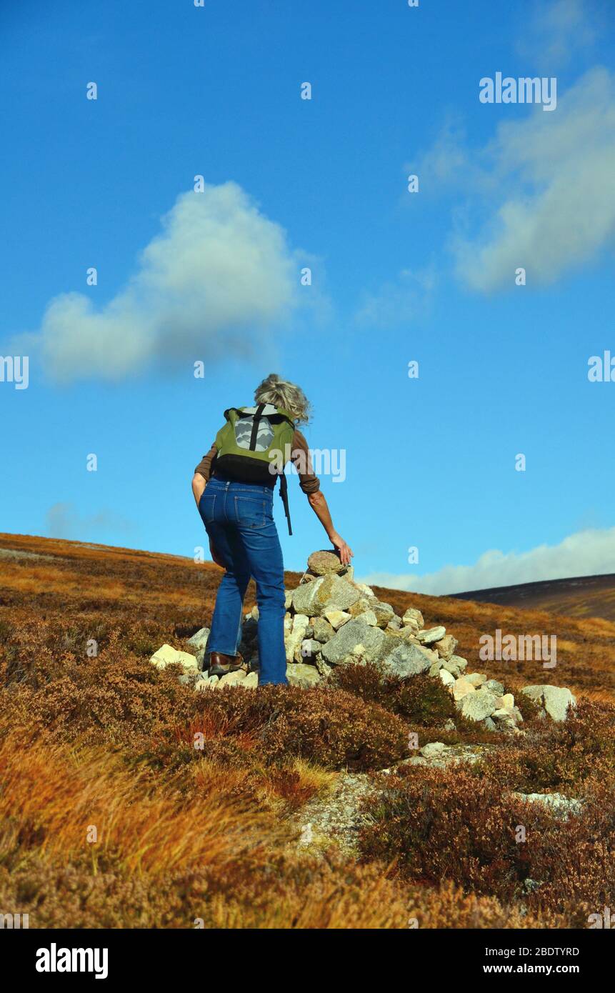A woman adding a rock to a cairn at the side of a footpath leading to the top of Mount Keen, at glen Esk Stock Photo