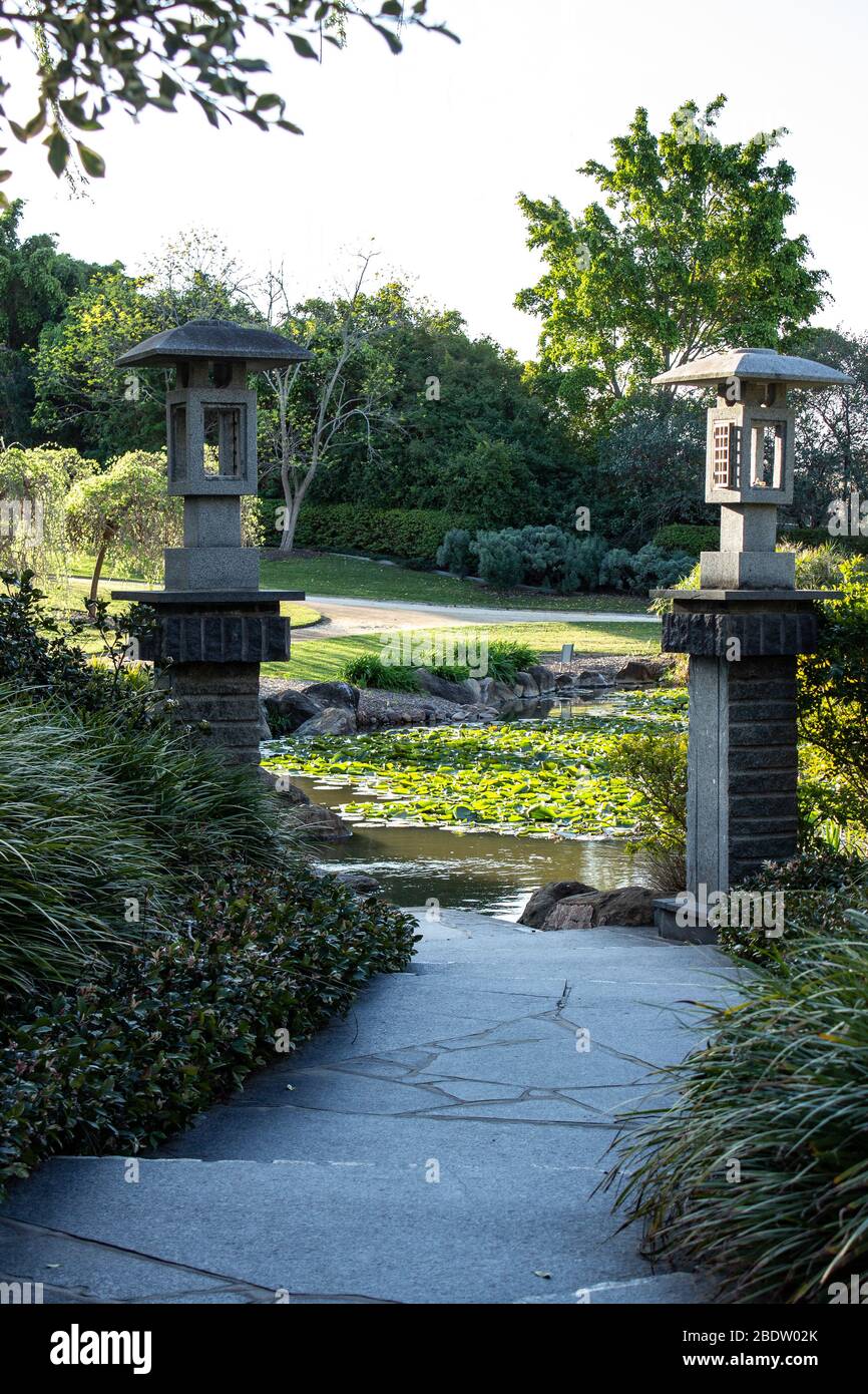 Winding garden path with Chinese style lanterns on pillars leading to water lilies floating on pond with trees in background Stock Photo