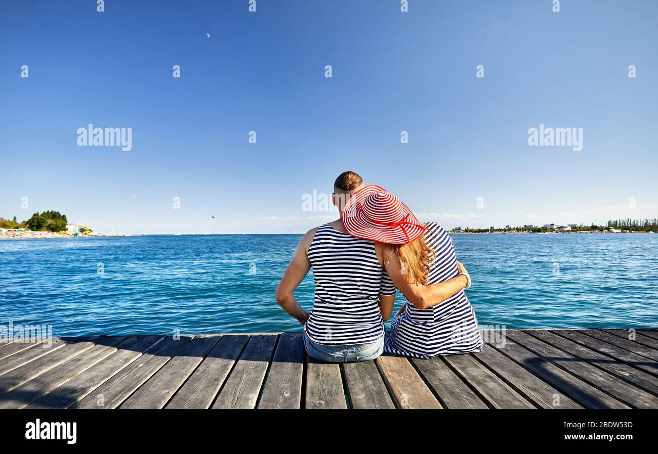 Romantic couple in striped dress sitting on pier and looking at blue lake Issyk Kul in Kyrgyzstan Stock Photo