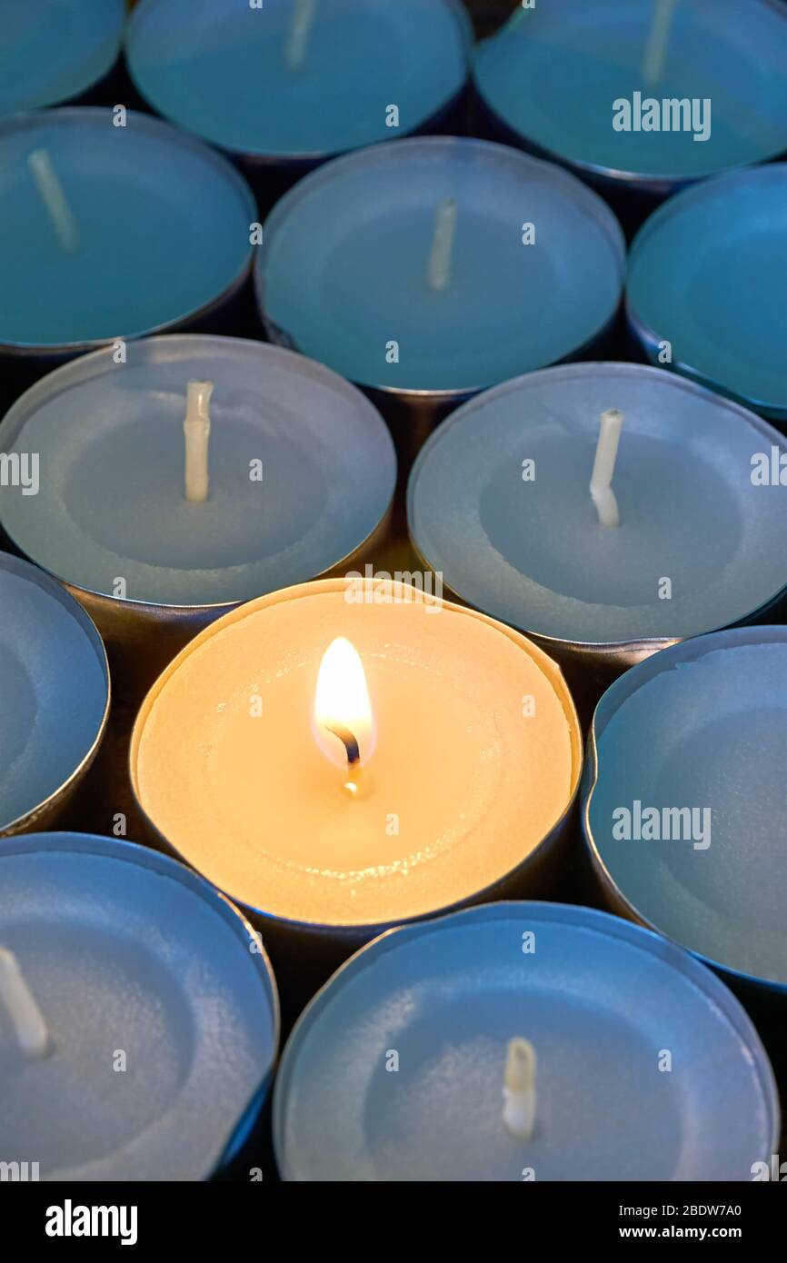 Closeup of tea lights with one being lit. A single warm orange flame between candles in blue shadow. Christmas, mourning or condolence concept. Stock Photo